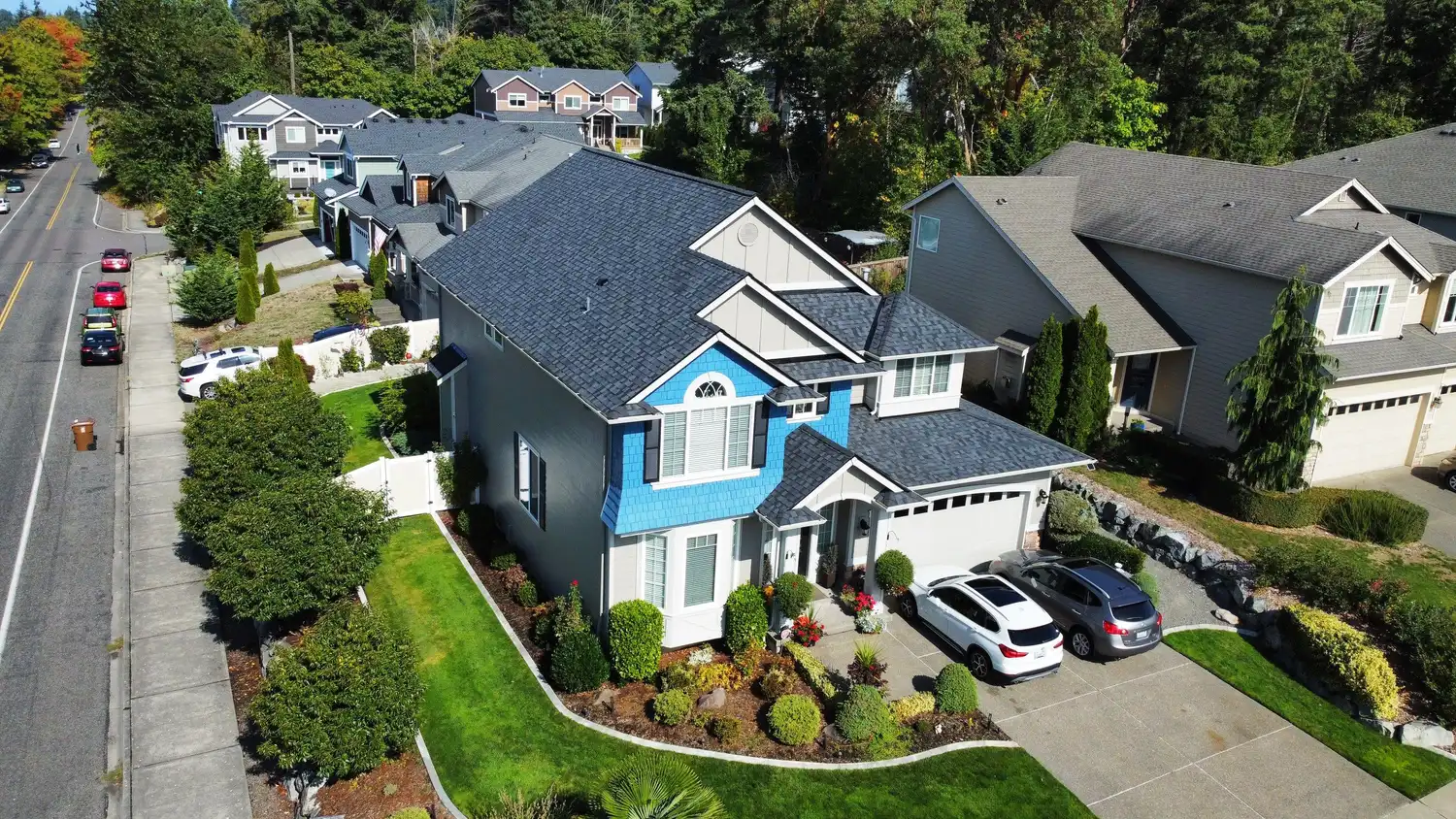 Aerial view of a suburban neighborhood with houses, one prominently featuring blue accents, a manicured lawn, and parked cars.