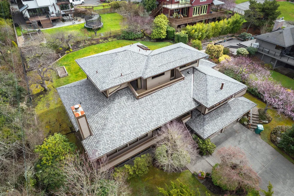 Aerial view of a large, multi-level residential home with a gray shingle roof surrounded by trees and landscaped gardens.