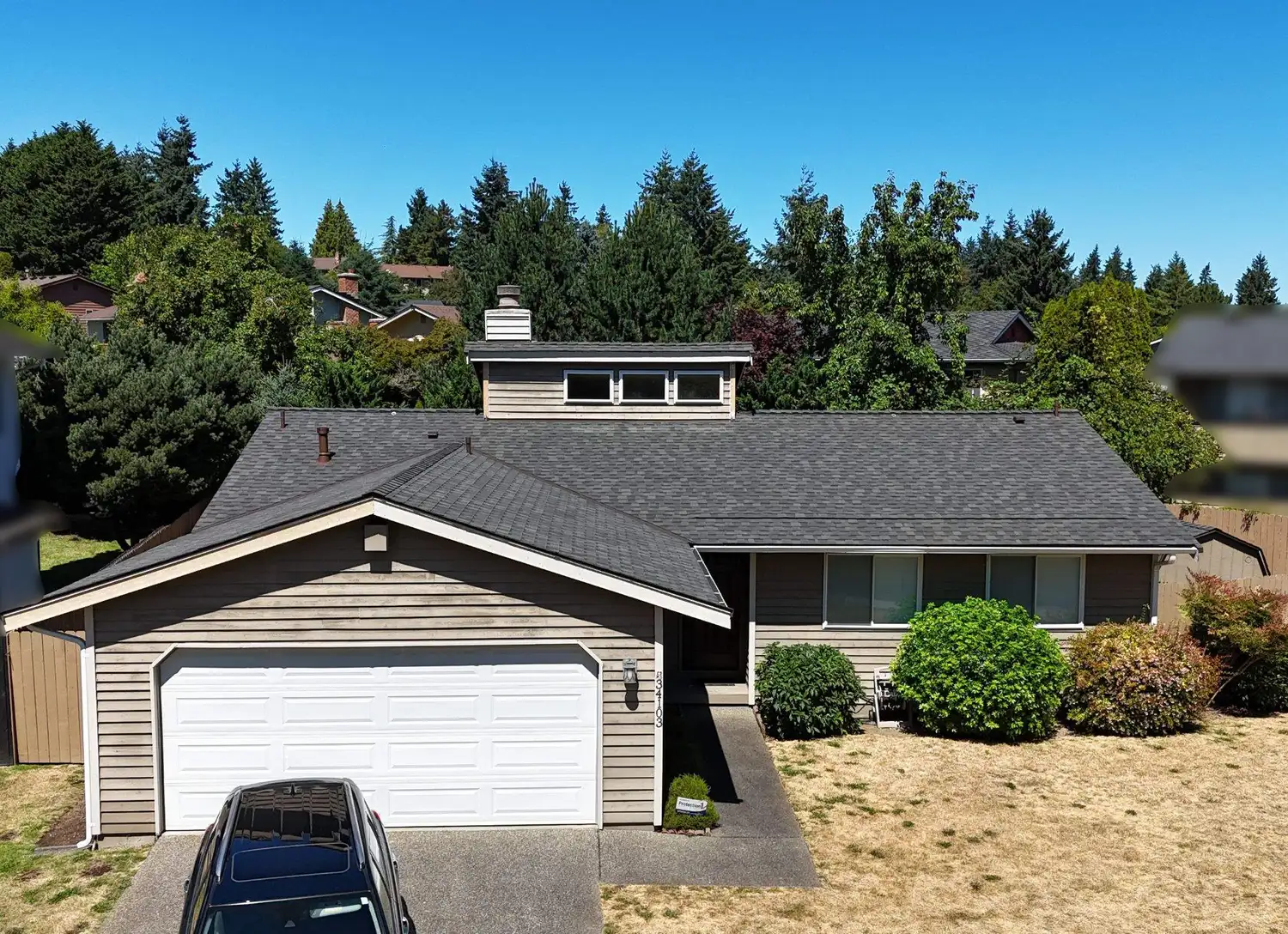 A single-story house with a gray roof, light brown siding, and a white garage door, surrounded by greenery and a clear blue sky.