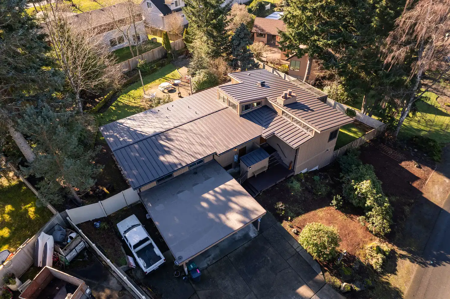Aerial view of a house surrounded by trees and greenery, with a driveway and neighboring homes visible.