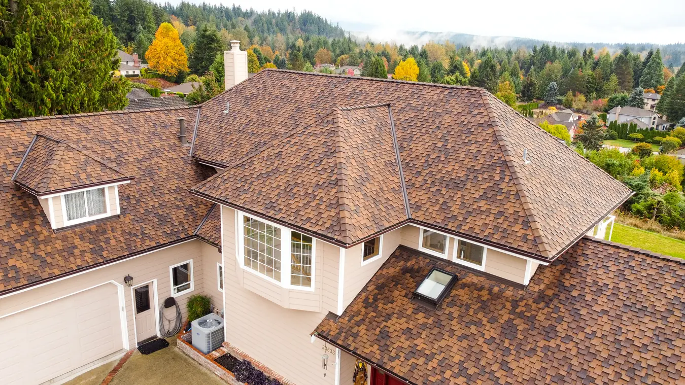 Aerial view of a suburban house with a brown shingle roof, surrounded by trees and hills in the background.