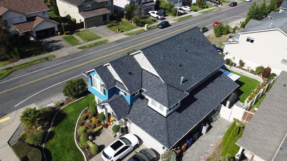 Aerial view of a residential neighborhood featuring a house with a dark roof, landscaped yard, and parked cars on the street nearby.