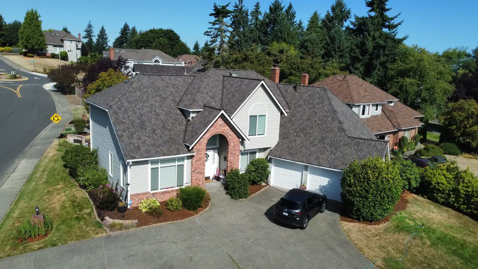 Aerial view of a suburban house with a gray roof and brick accents, surrounded by trees and a well-maintained lawn, with a driveway and parked car.