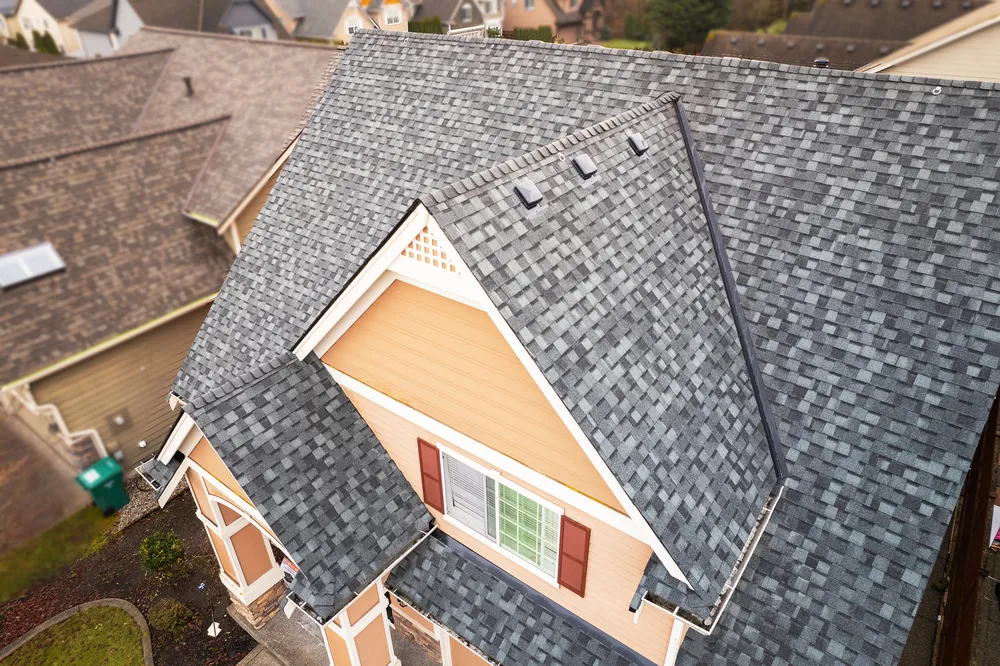 Aerial view of a house with a gray shingle roof and light-colored siding, surrounded by other homes.