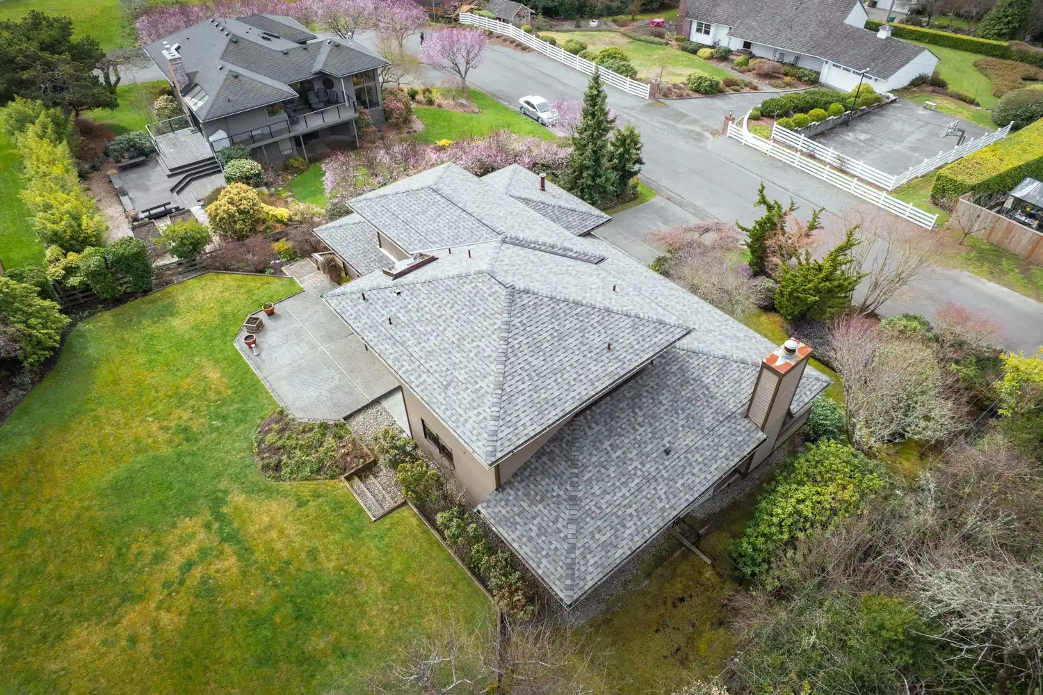 Aerial view of a large house with a grey roof, surrounded by greenery and trees, with another house visible in the background.