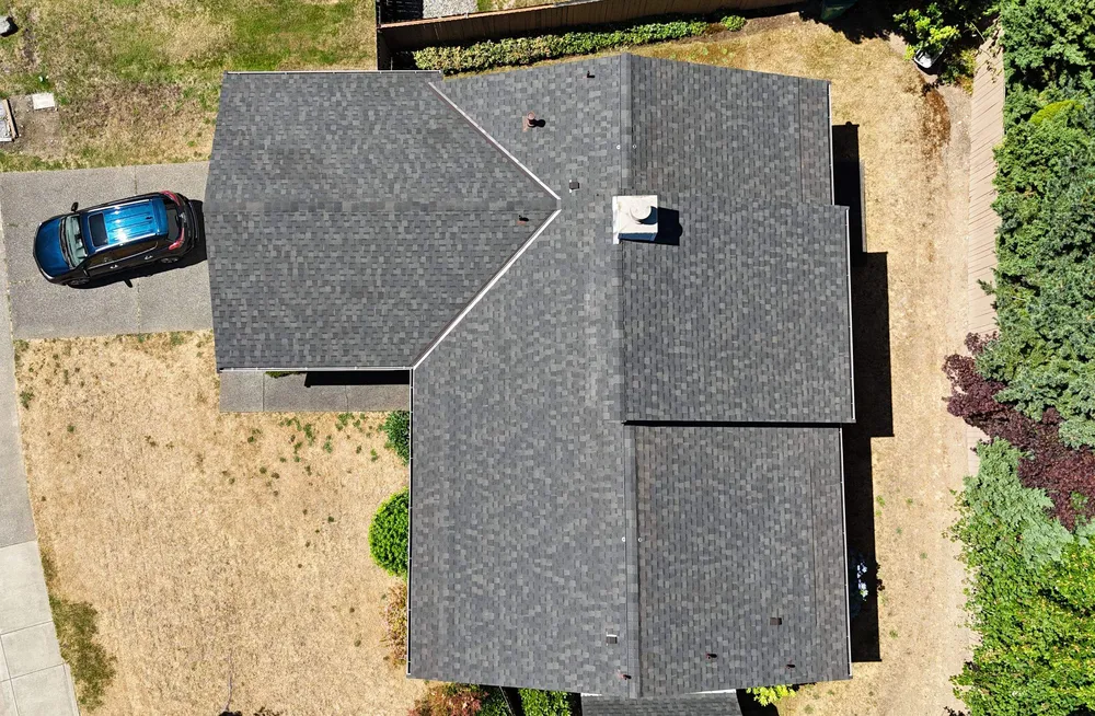 Aerial view of a house with a dark roof and a car parked in the driveway, surrounded by grass and shrubs.