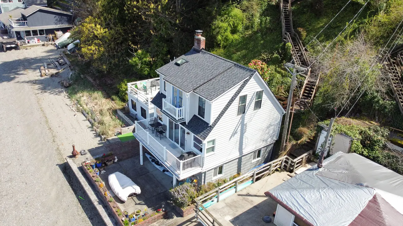Aerial view of a beachside house with a gray roof, balcony, and surrounding landscape.