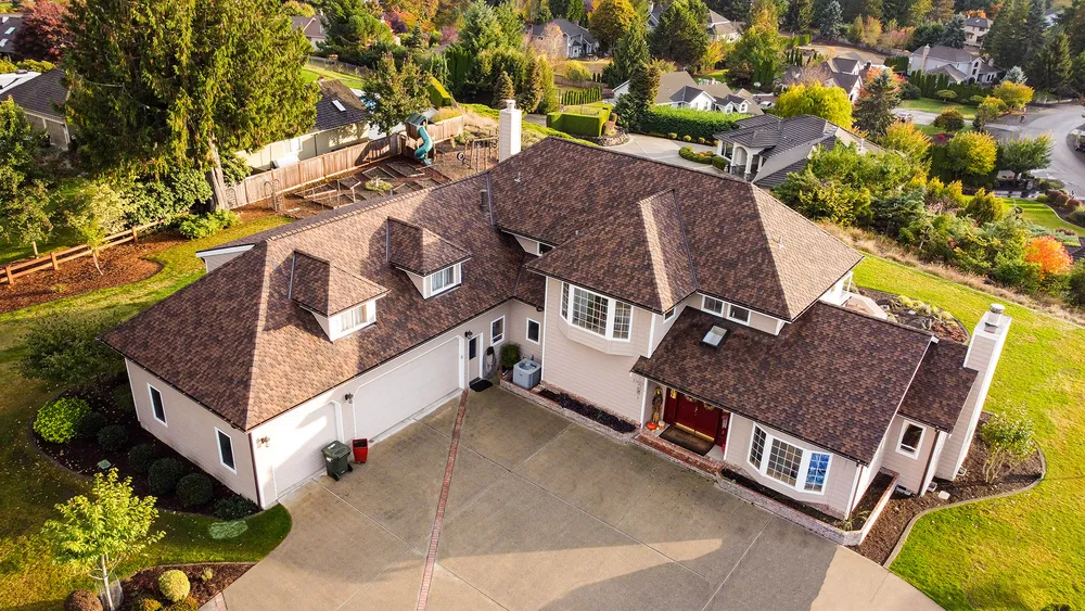 Aerial view of a large house with a brown roof, surrounded by greenery and trees in a suburban area.