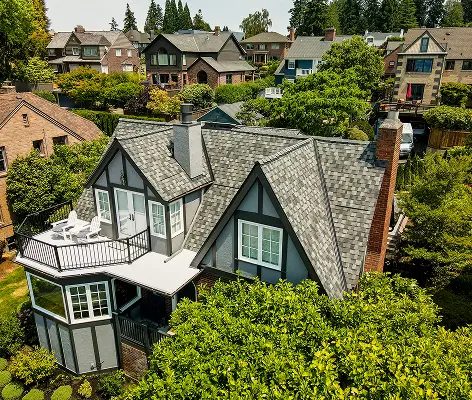 Aerial view of a gray house with a pitched roof, surrounded by trees and other houses in a residential area. The house features a balcony and a well-maintained garden.