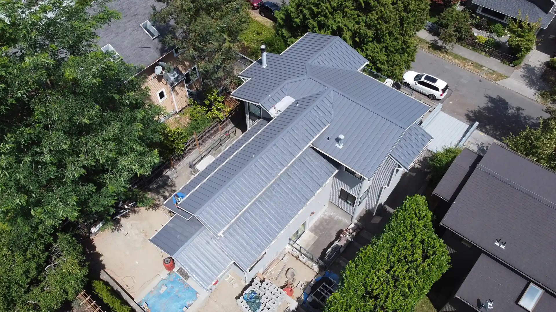 Aerial view of a modern single-story home with a gray metal roof, surrounded by trees and a paved driveway.