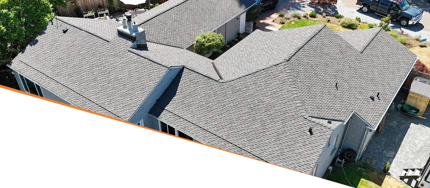 Aerial view of a residential roof with multiple slopes and shingles, surrounded by a yard and driveway.