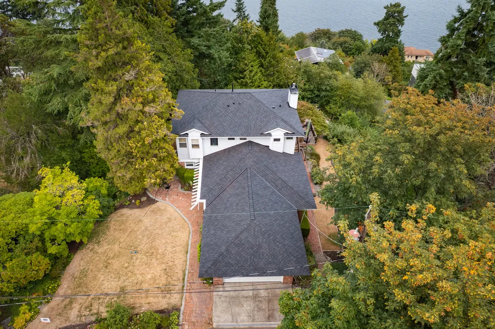 An aerial view of a large, two-story house surrounded by trees and greenery, with a landscaped yard and a lake in the background.