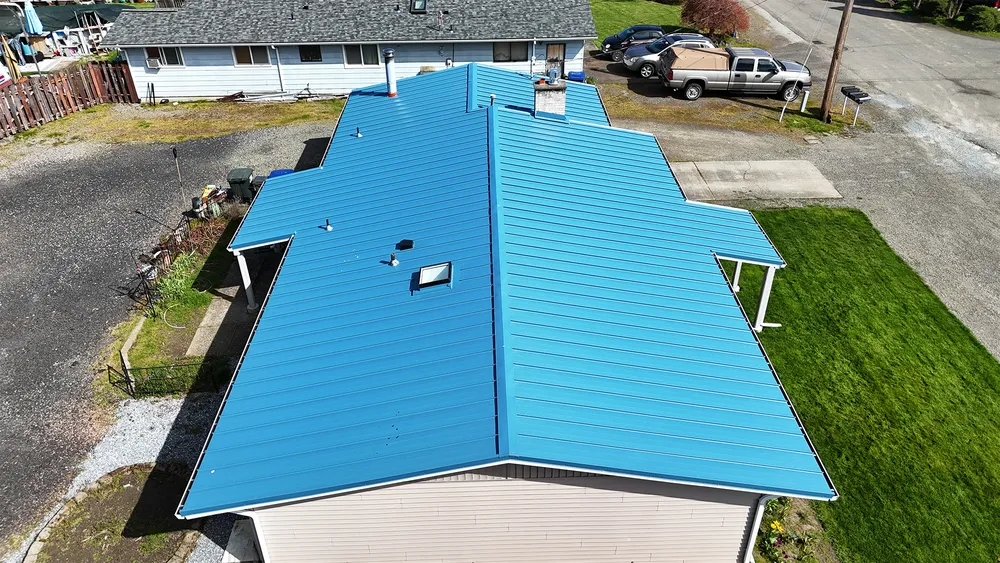 Aerial view of a house with a blue metal roof, surrounded by green grass and a gravel driveway.