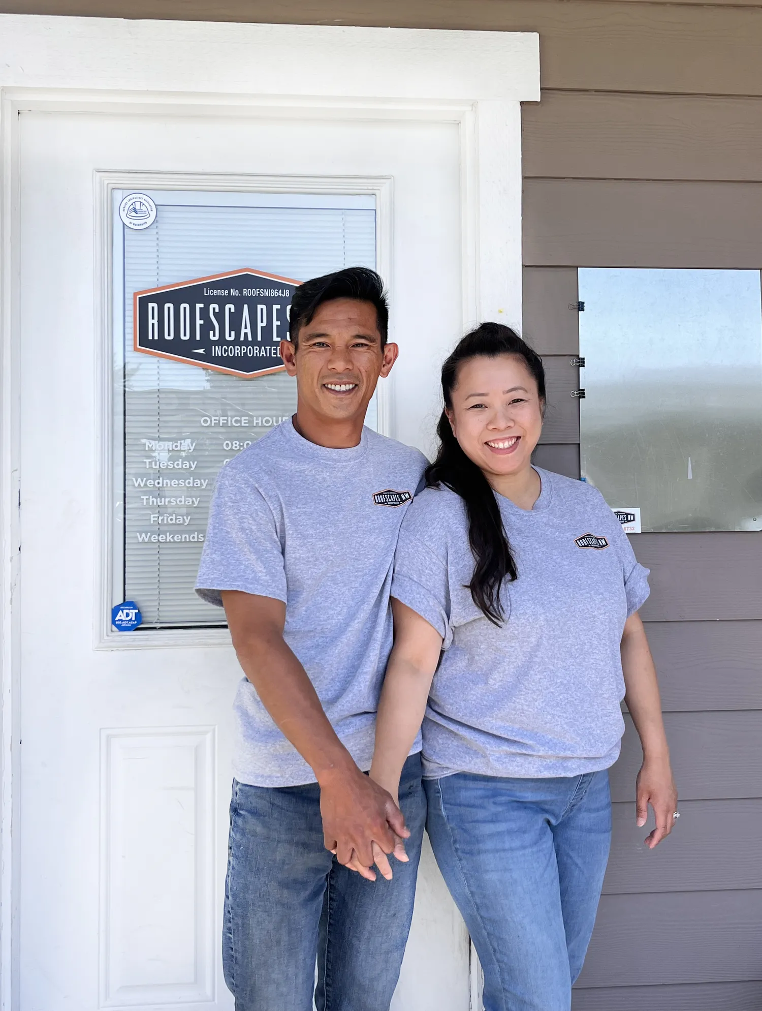 A young man and woman, both wearing gray t-shirts, stand in front of a door with a sign that reads "Roofscapers Incorporated." They are smiling and holding hands.
