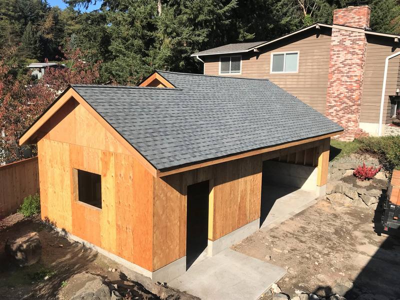 A newly constructed shed or small building with wooden walls and a gray shingle roof, situated in a backyard among trees and near a house.