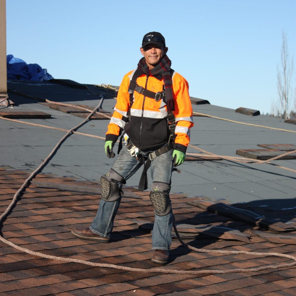 A person wearing a bright orange jacket and safety gear stands on a roof, with ropes and roofing materials around.