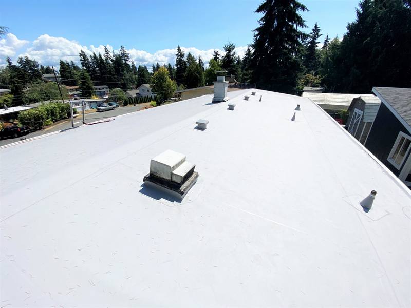 A flat roof with several vents and chimneys, surrounded by trees and buildings under a clear blue sky.