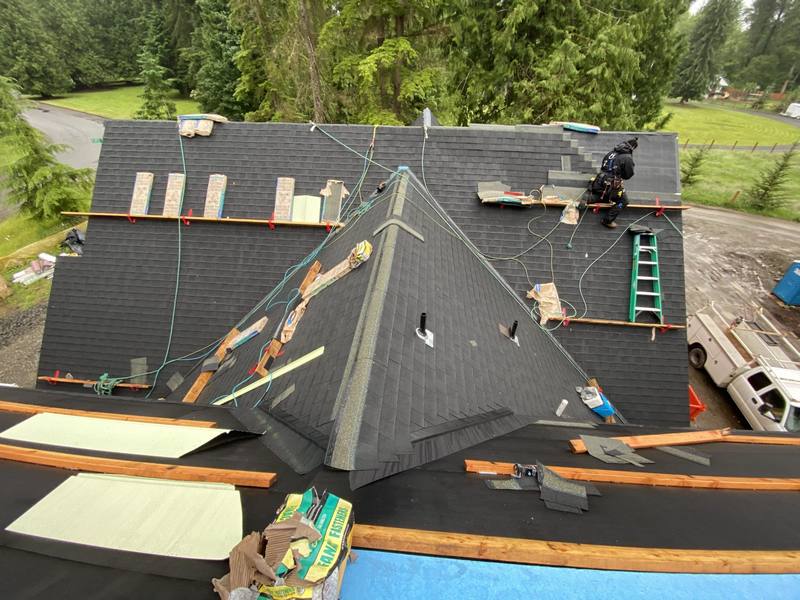 Aerial view of a roofing project, showing workers installing shingles on a sloped roof, with construction materials and ladders visible.