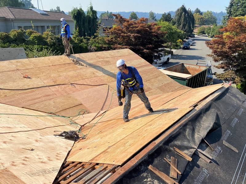 Two workers wearing hard hats are installing roofing materials on a sloped roof, with trees and houses visible in the background.