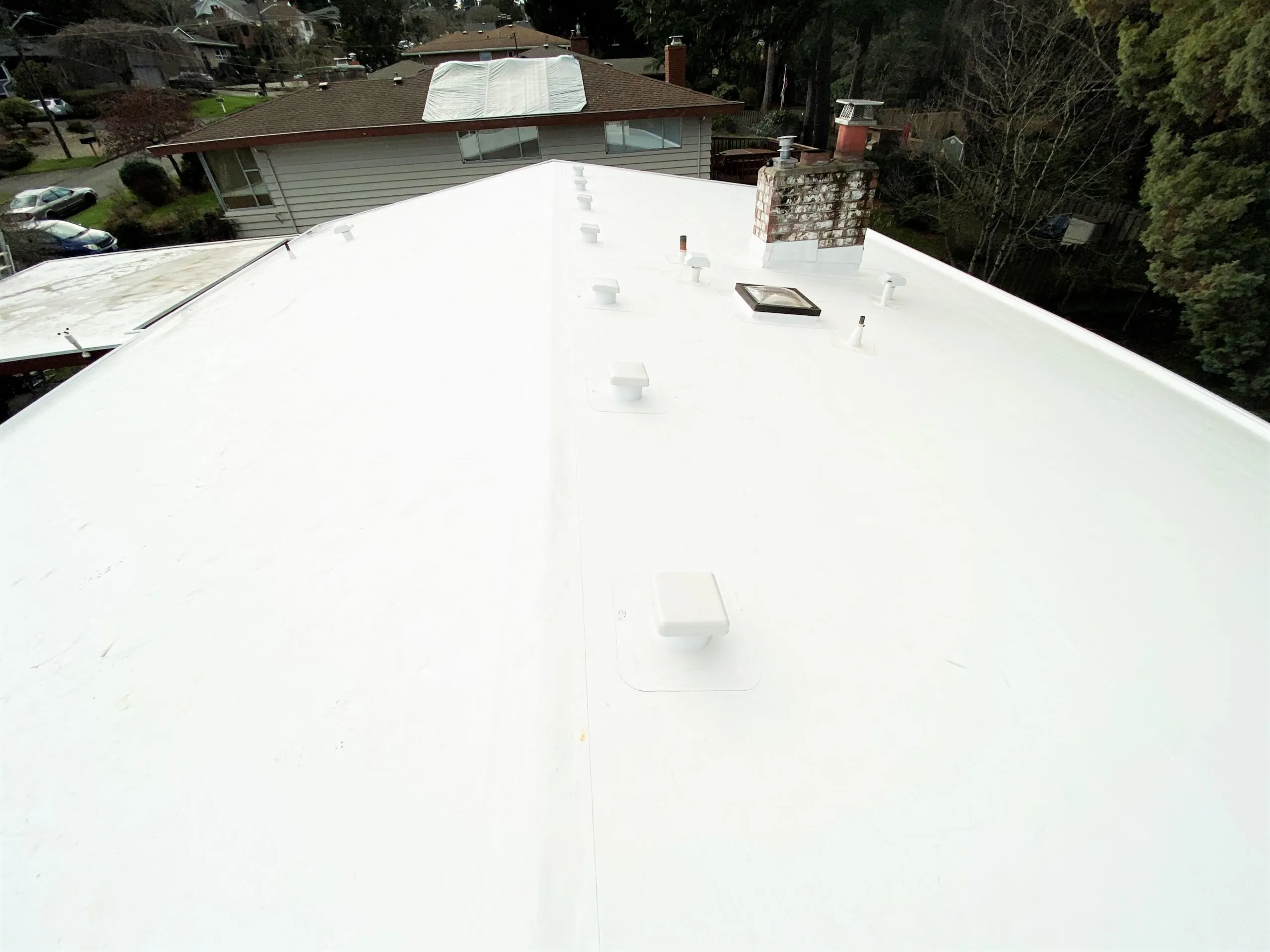 Aerial view of a white roof with ventilation pipes and a chimney, surrounded by trees and houses.