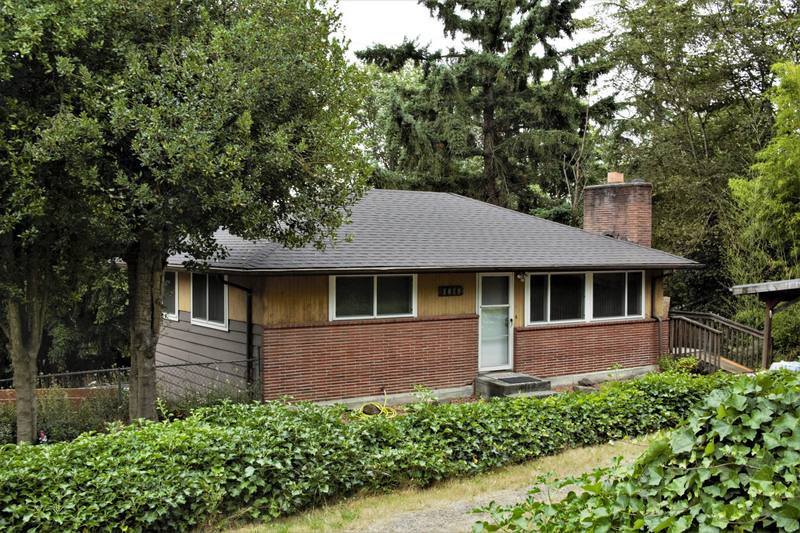 A mid-century modern house with a brick exterior, large windows, and a sloped roof, surrounded by greenery and trees.