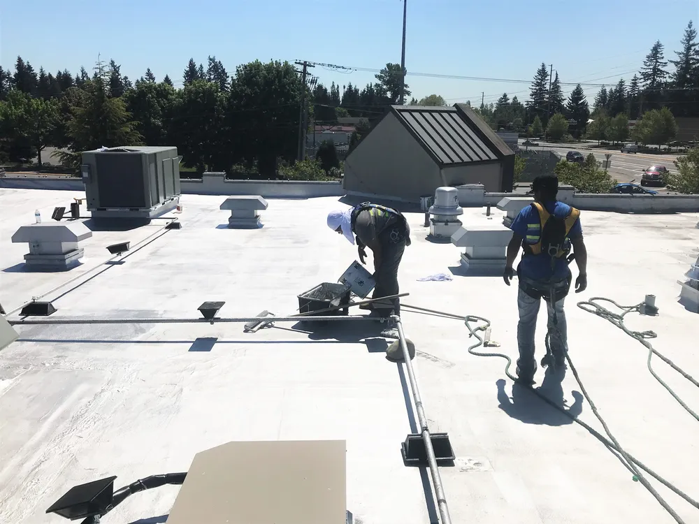 Two workers on a flat roof, one kneeling and the other standing, with various tools and rooftop equipment visible.