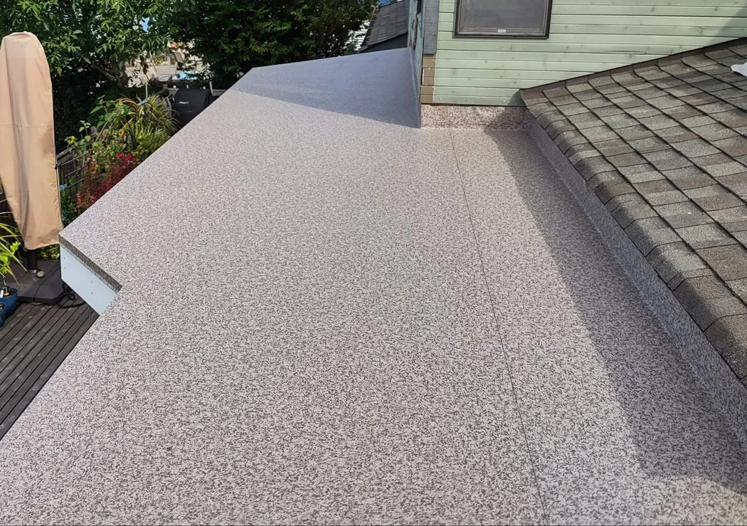 A flat roof with a smooth, textured surface, transitioning from one section to another. One side features a sloped roof with shingles. Surrounding greenery is visible in the background.